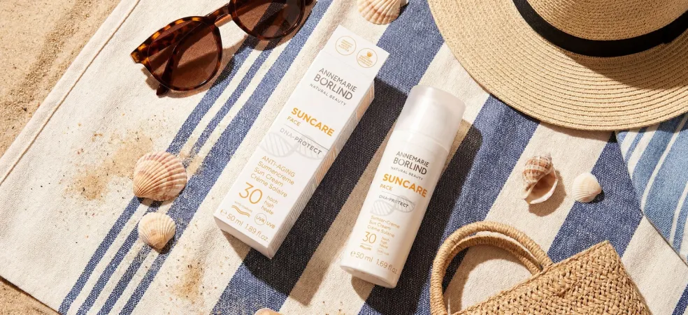 Opaľovací anti-aging krém ANNEMARIE BÖRLIND DNA Protect SPF 30 na plážovom uteráku medzi slnečnými okuliarmi, slamákom a mušličkami – ochrana pleti pred slnkom