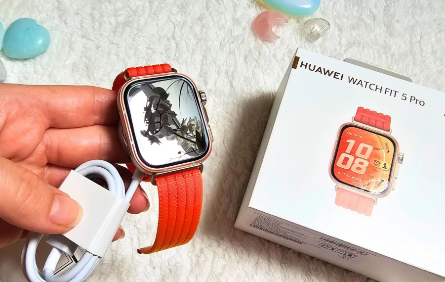 Huawei Watch Fit 5 Pro NFC v oranžovej farbe v otvorenom balení, produkt držiaci v ruke