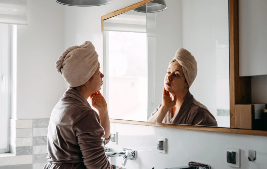 Žena v župane si pred zrkadlom kontroluje stav pleti počas skincare rutiny