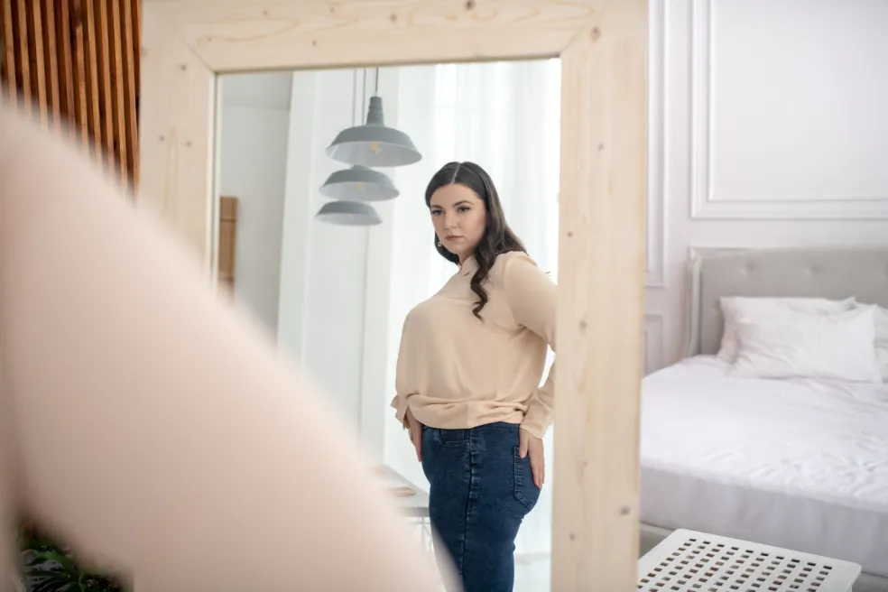 Mladá žena si so znepokojeným výrazom prezerá svoju postavu v zrkadle