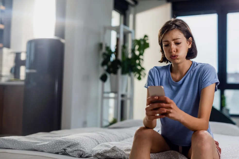 Mladá žena sedí sama v spálni a znepokojene sleduje obsah v mobilnom telefóne