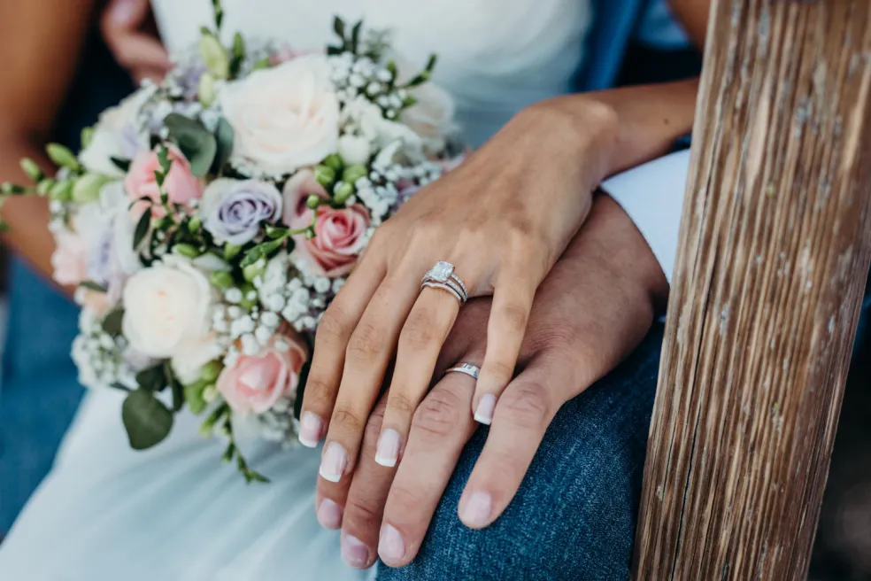 Ženská ruka s elegantne vrstvenými prsteňmi položená na manželovej ruke pri svadobnej kytiici