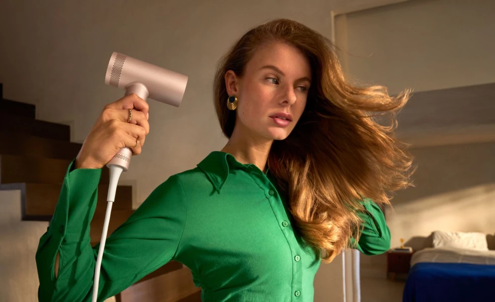 Žena v letnom outfite, ktorá si upravuje vlasy fénom alebo kulmou Philips v svetlej kúpeľni, s ďalšími beauty prístrojmi Philips položenými na kozmetickom stolíku.