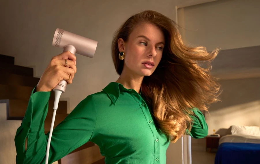 Žena v letnom outfite, ktorá si upravuje vlasy fénom alebo kulmou Philips v svetlej kúpeľni, s ďalšími beauty prístrojmi Philips položenými na kozmetickom stolíku.