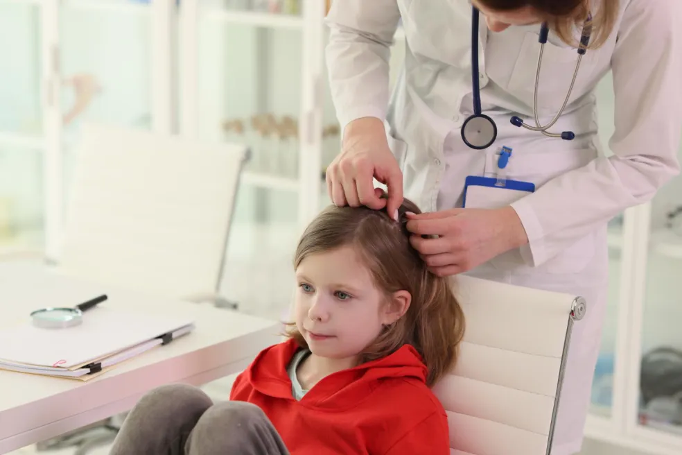 Pediatrička v ambulancii kontroluje školáčke pokožku hlavy a vlasy pri podozrení na vši, zatiaľ čo mama sedí vedľa