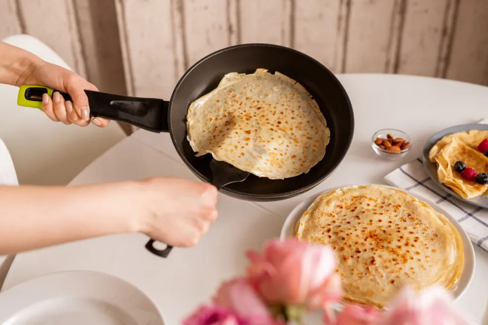 Ruky mladej ženy držiace panvicu s horúcou palacinkou počas prípravy v kuchyni
