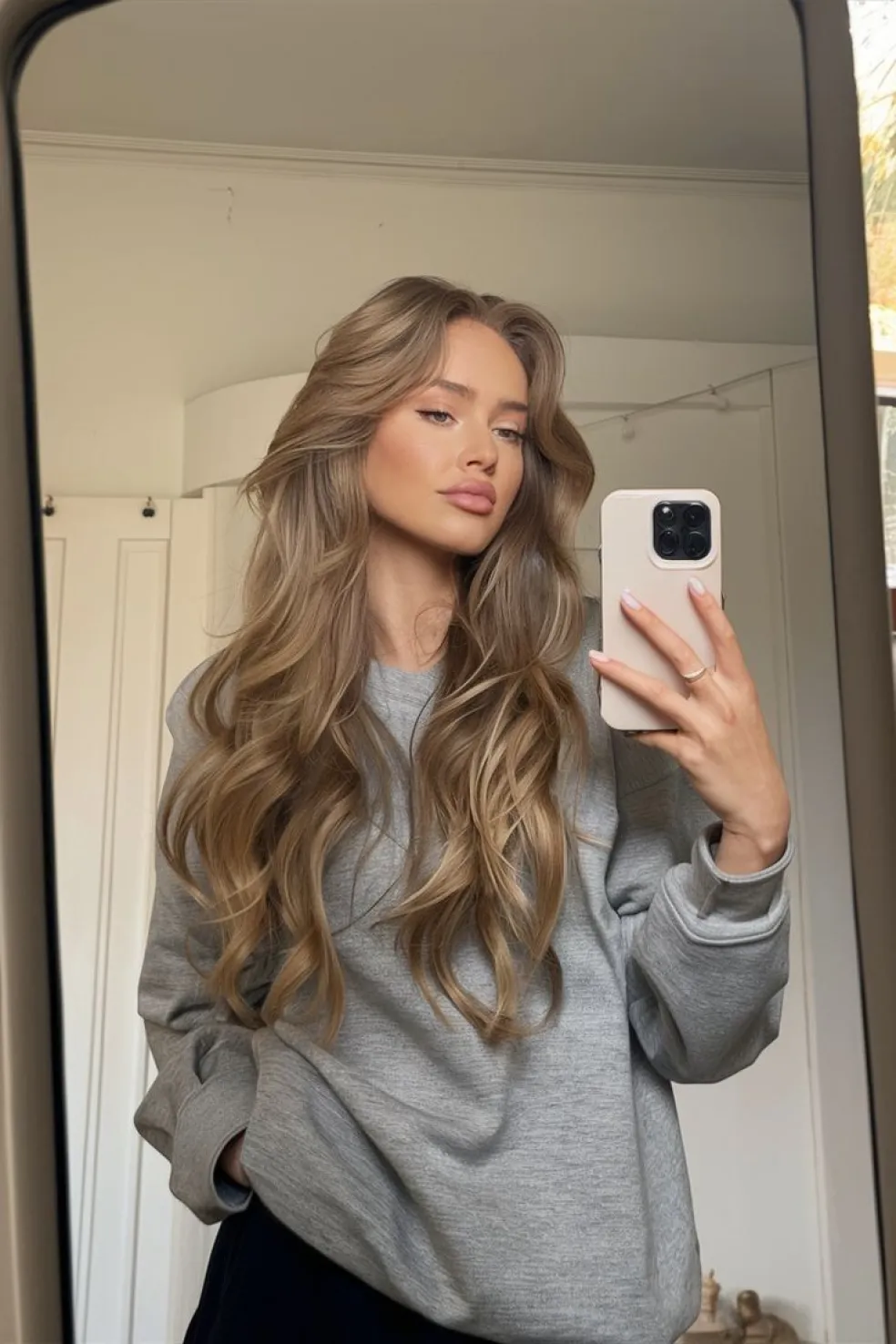 Žena s dlhými vlnitými vlasmi v béžovo-hnedom odtieni s jemnými blond odleskami si fotí selfie v zrkadle