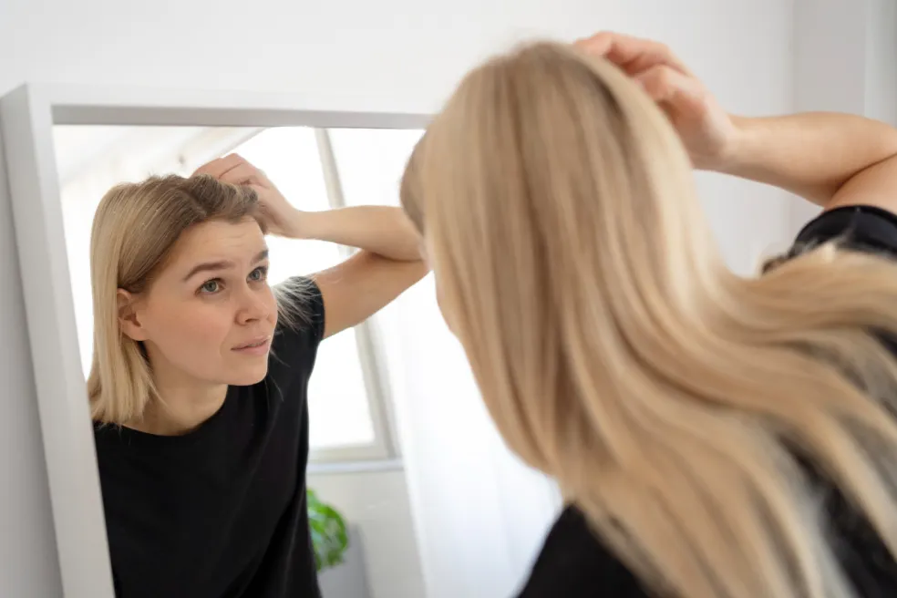 Žena si pred zrkadlom kontroluje pokožku hlavy a vlasové korienky