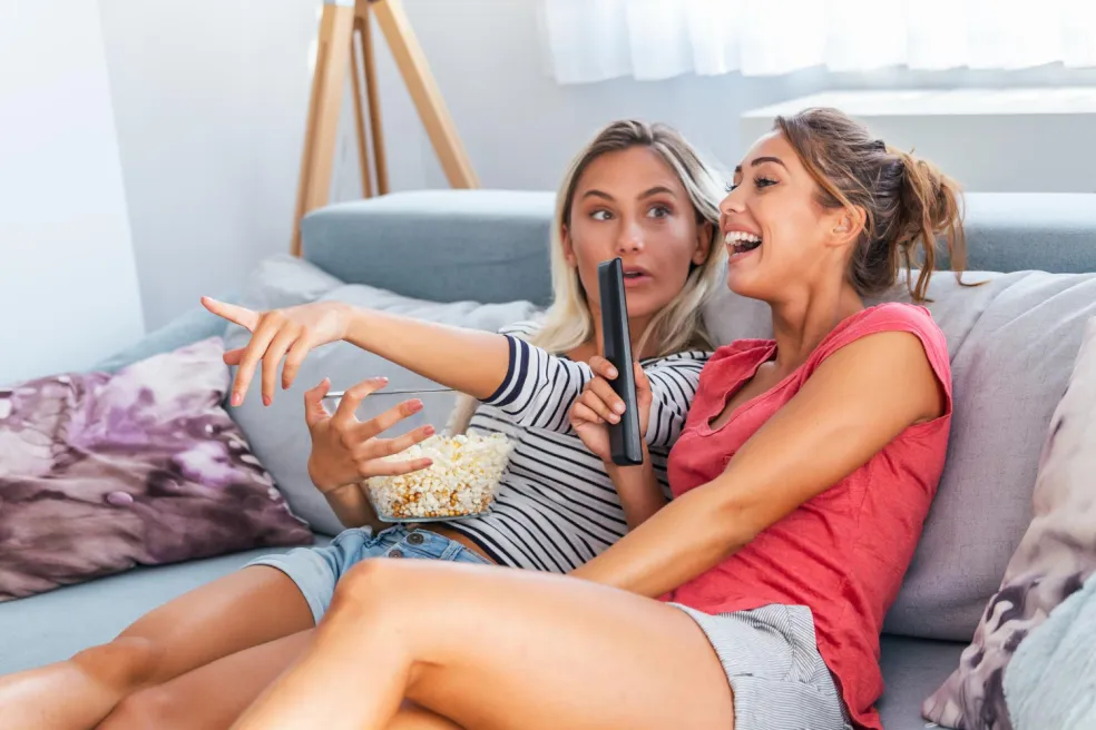 Dve kamarátky sledujú komédiu v televízii, smejú sa na gauči a jedia popcorn