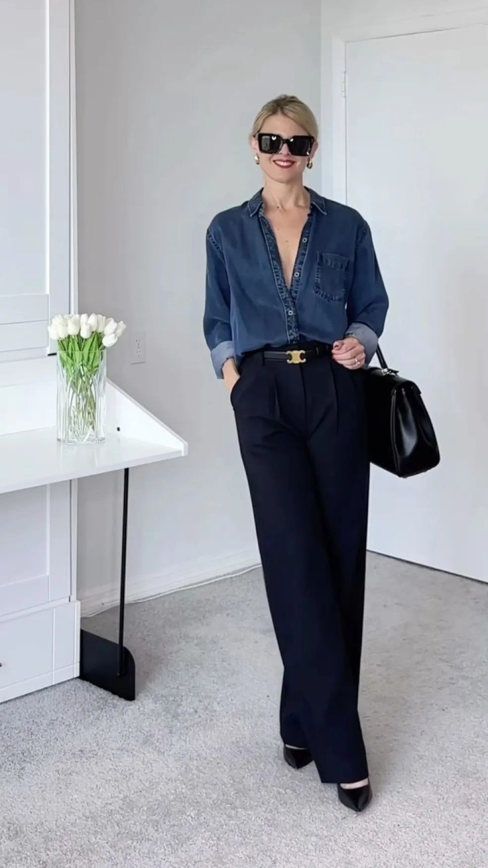Elegantný outfit s tmavomodrou košeľou zastrčenou do širokých čiernych nohavíc, bordovou kabelkou, zlatými náušnicami a čiernymi sandálmi