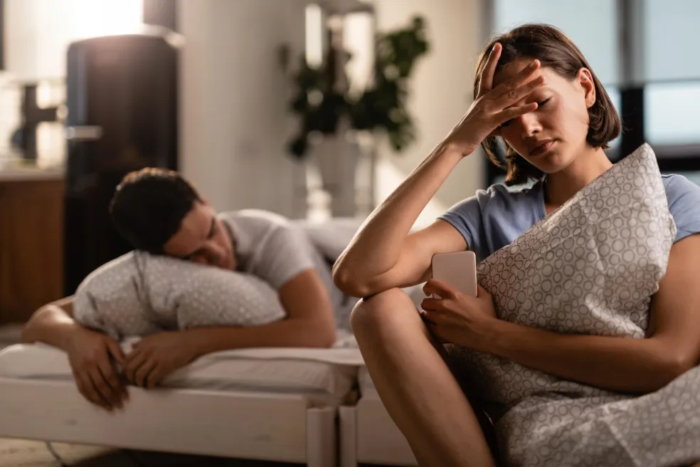 Emocionálne vyčerpaná žena sedí s rukou na čele, zatiaľ čo jej partner leží v pozadí na pohovke. Fotografia ilustruje frustráciu, partnerské problémy a únavu z čakania na zmenu vo vzťahu