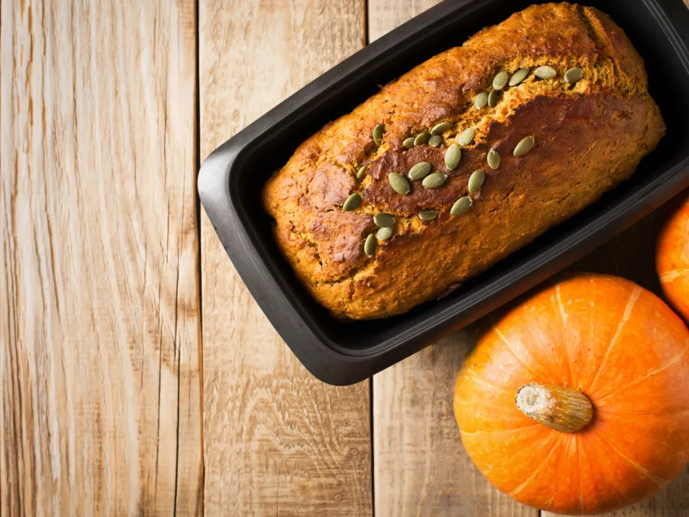 Upečený tekvicový banana bread so zlatohnedou kôrkou a tekvicovými semienkami v podlhovastej forme na pečenie, s čerstvými tekvicami na drevenom stole