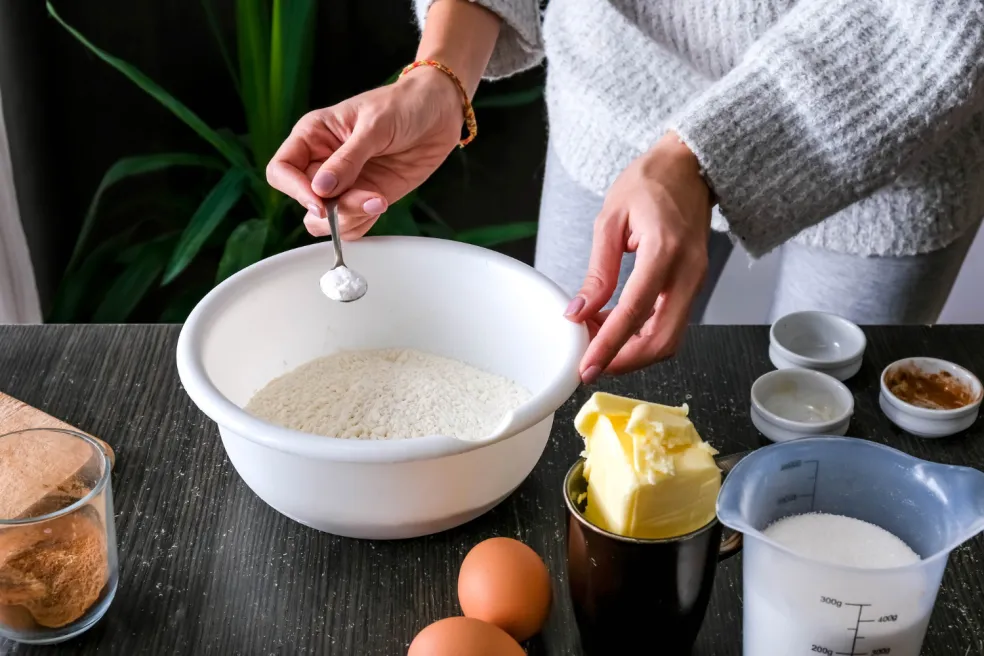 Príprava cesta na tekvicový banana bread – pridávanie kypriacej prísady do múky v mise, s maslom, vajíčkami, cukrom a škoricou na pracovnej doske