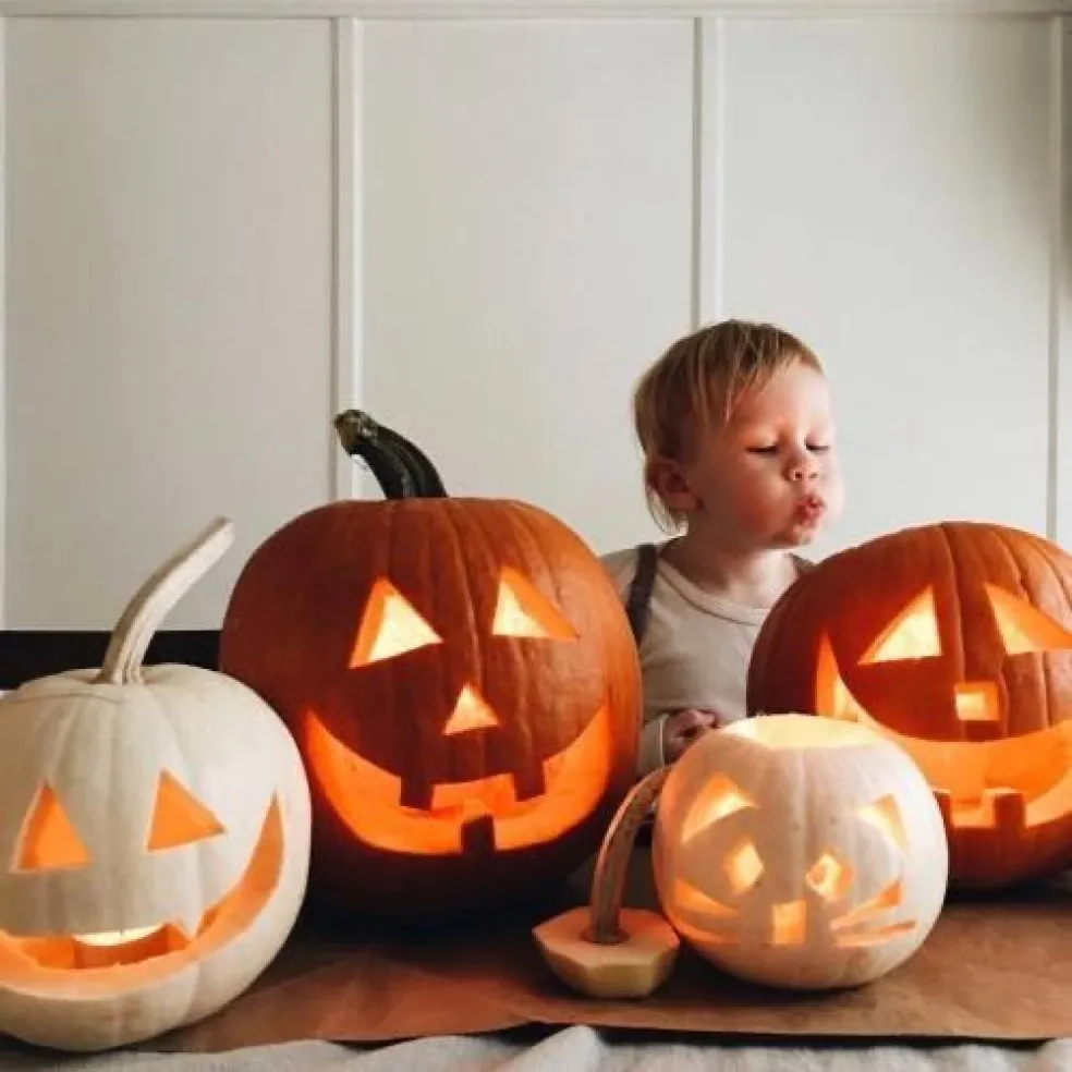 Dieťa za stolom so štyrmi svietiacimi vyrezávanými halloweenskymi tekvicami rôznych veľkostí, s veselými tvárami a strašidelnými úsmevmi