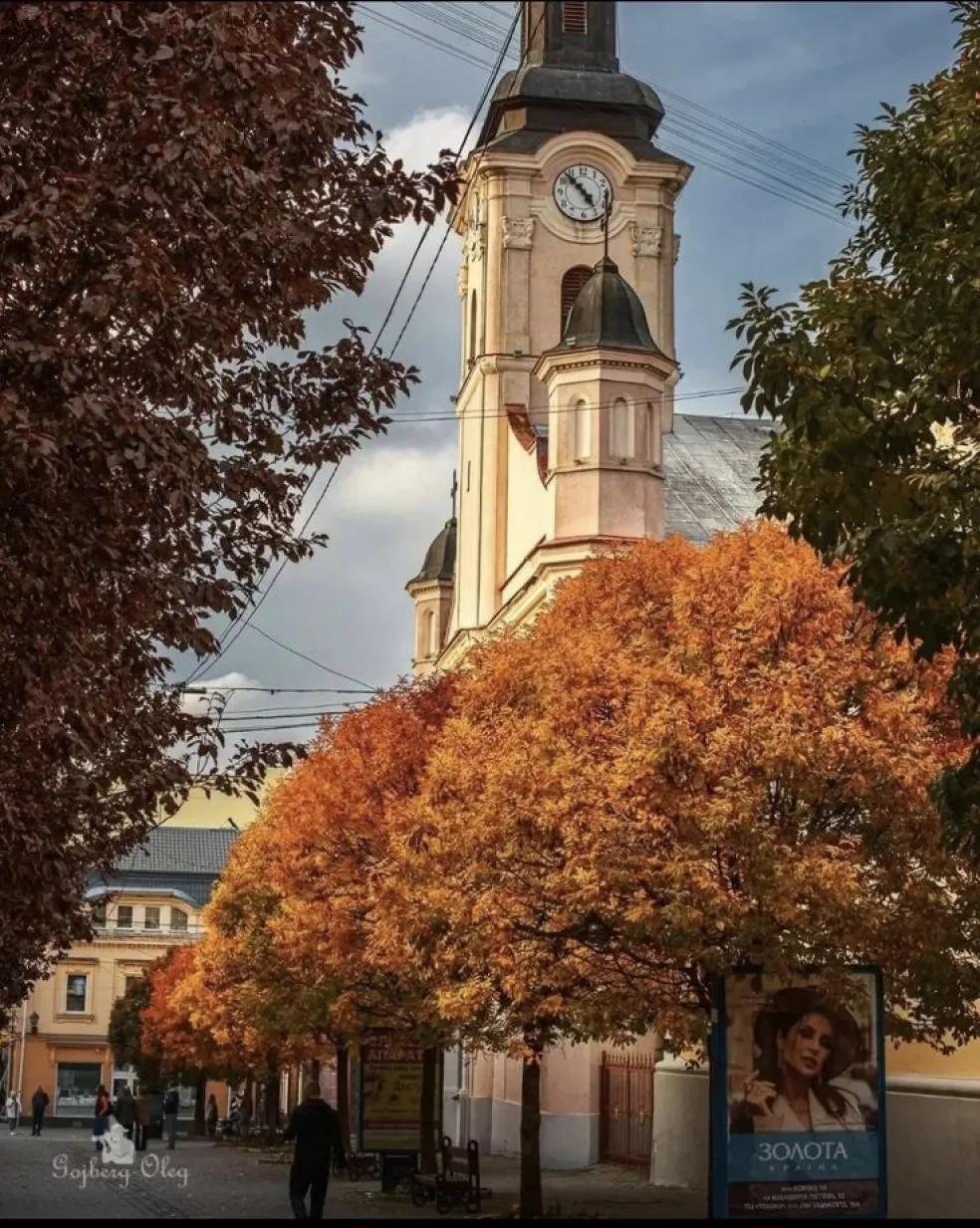 Jesenná atmosféra historického centra mesta s kostolnou vežou a hodinami, obklopenou stromami so žiarivým oranžovým a červeným lístím