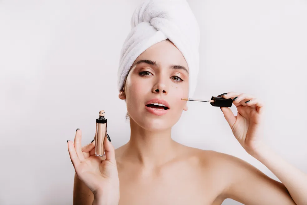 Žena s uterákom na hlave nanáša tekutú dekoratívnu kozmetiku aplikátorom na líce – príprava pleti a líčenie pre dlhotrvajúci make-up