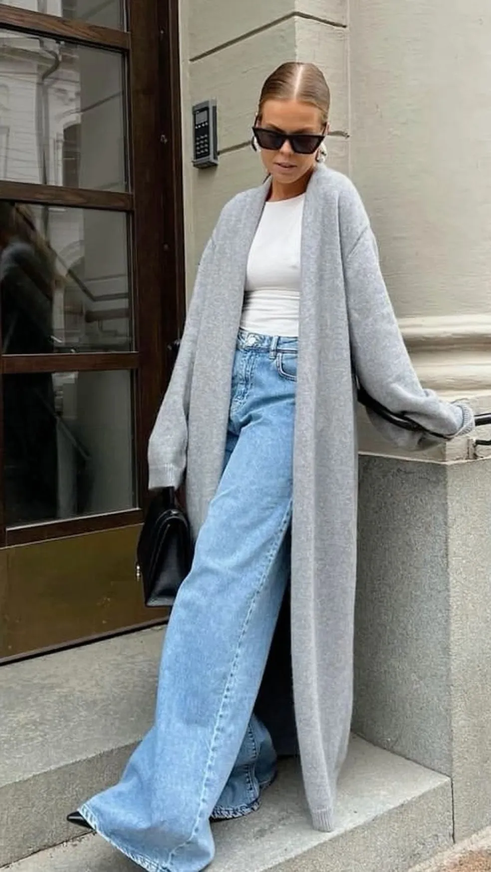 Žena v dlhom sivom maxi kardigáne, bielom tričku a svetlomodrých širokých džínsoch – pohodlný mestský outfit do práce