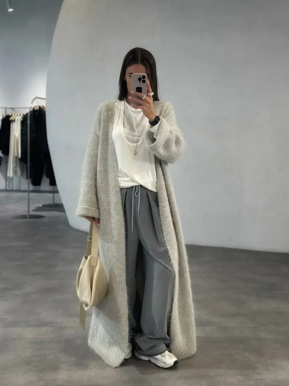 Žena v dlhom svetlobéžovom maxi kardigáne, bielom tričku, sivých voľných nohaviciach a teniskách – pohodlný víkendový outfit na jesennú prechádzku