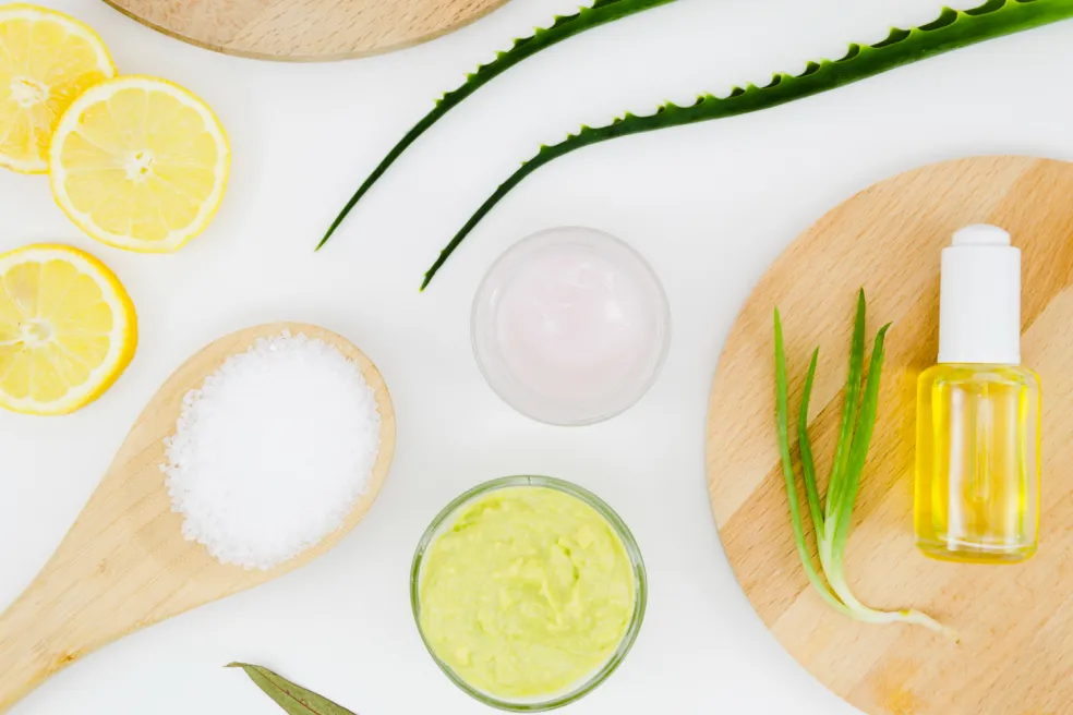 Pohľad zhora na citróny, listy aloe vera, misky s kozmetickými zmesami, drevenú lyžicu so soľou a fľaštičku oleja, znázorňujúce ingrediencie na domácu vlasovú masku