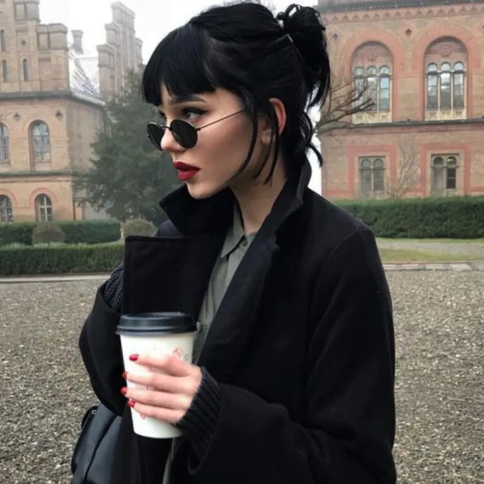 Žena s čiernymi vlasmi upravenými do ležérneho drdola s ofinou, v tmavých okuliaroch a čiernom kabáte – inšpirácia na elegantný účes s čiernou farbou vlasov