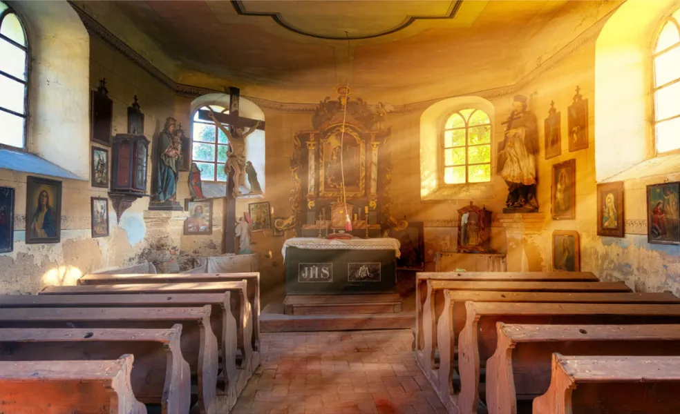 Interiér prázdneho historického kostola s drevenými lavicami, oltárom, krížom a náboženskými obrazmi, osvetlený slnečnými lúčmi. Ilustračná fotografia k symbolike sna o prázdnom kostole